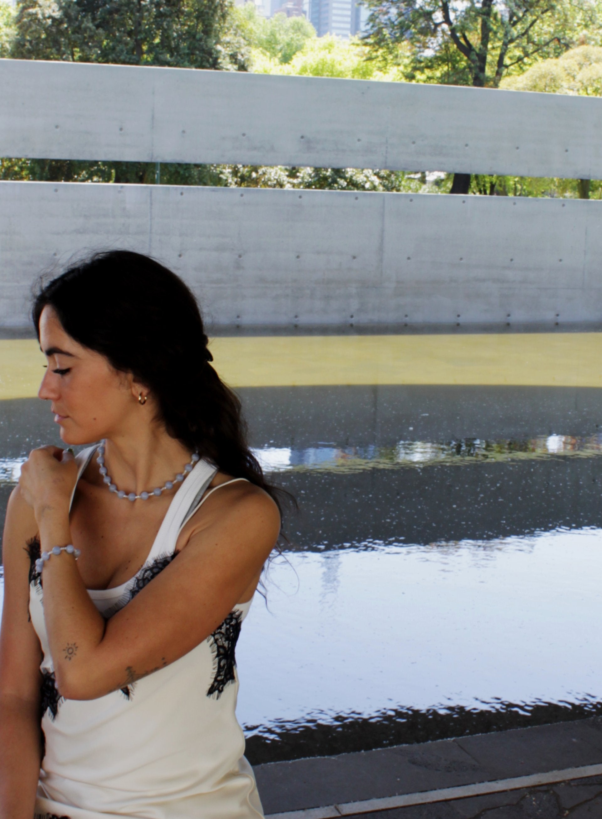 Woman sitting on a bench by a waterfront with trees and buildings in the background with blue stones bracelet and necklace