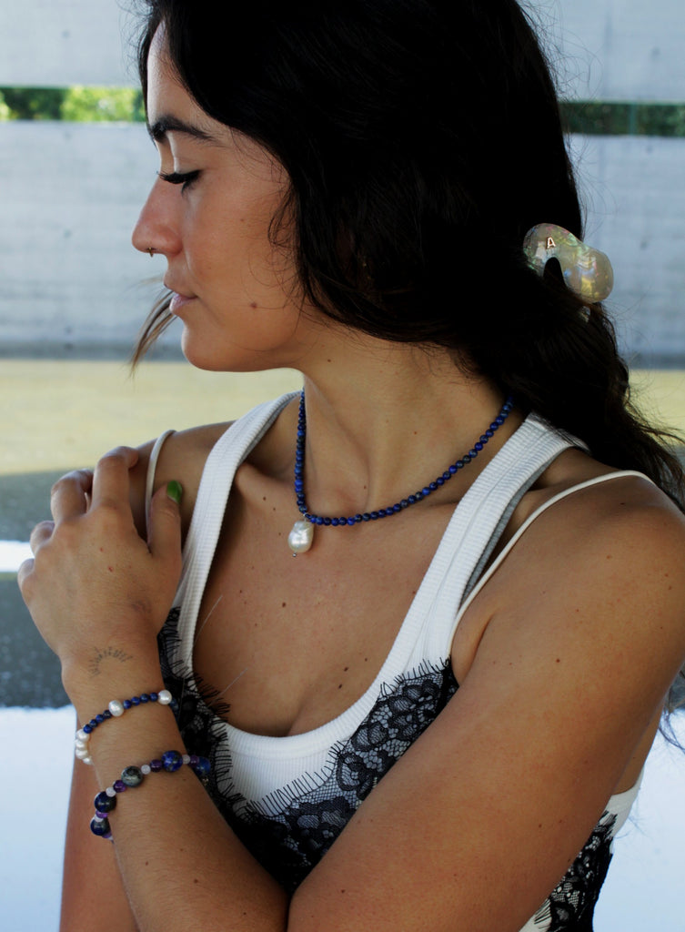 Woman with a ponytail wearing a blue beaded necklace and baraoque pearl charm and white top.