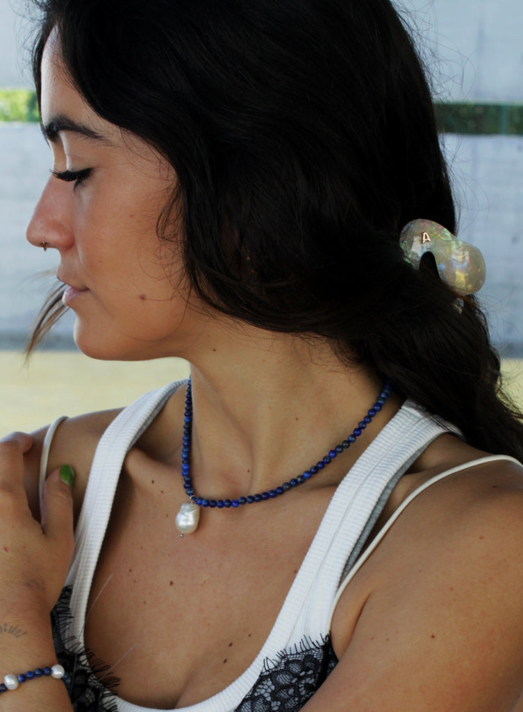 Woman with a ponytail wearing a blue beaded necklace and baraoque pearl charm and white top.