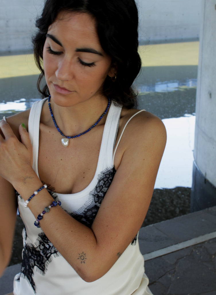 Woman with a ponytail wearing a blue beaded necklace and baraoque pearl charm and white top.