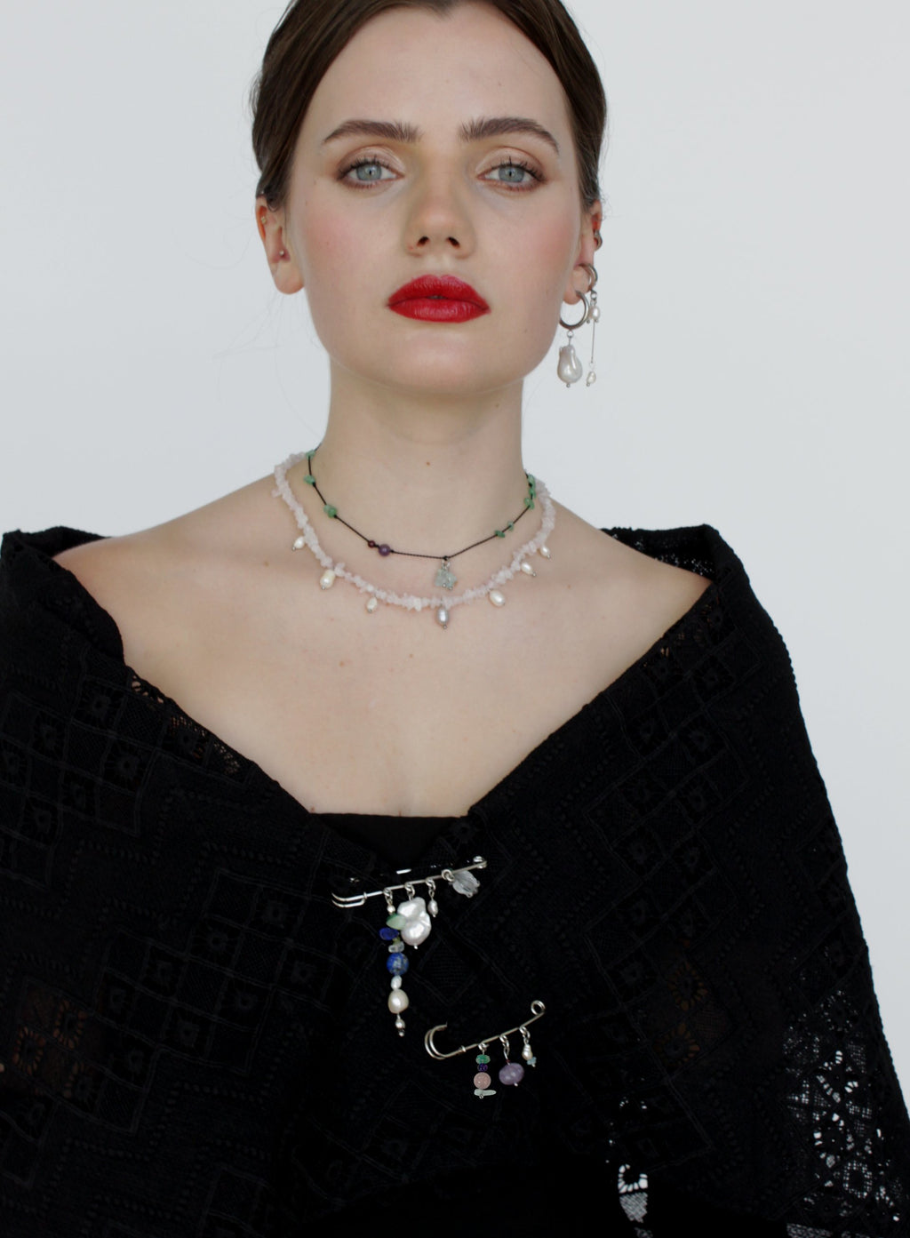 Woman wearing a black top with layered necklaces and pin brooches against a plain background